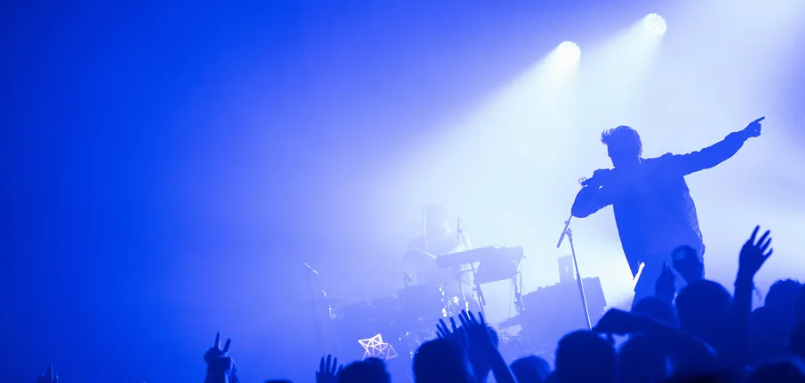 A silhouetted performer on stage, holding a microphone and engaging with an enthusiastic audience. The scene is illuminated by bright blue and white stage lights, creating a lively concert atmosphere with raised hands from the crowd.